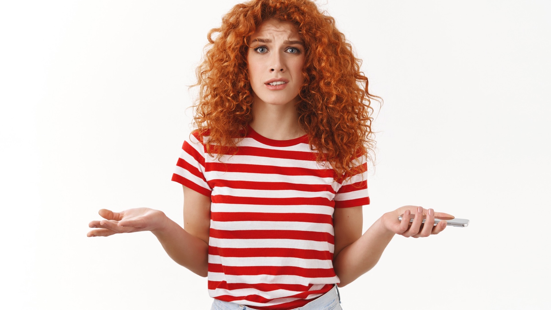 The image shows a young woman with curly red hair wearing a red and white striped t-shirt. She has a confused or questioning expression on her face, with her eyebrows raised and her mouth slightly open. She is holding a smartphone in one hand while her other hand is raised in a gesture of uncertainty or confusion, as if she is unsure or asking a question. The background is plain and white, drawing focus to her expression and body language.