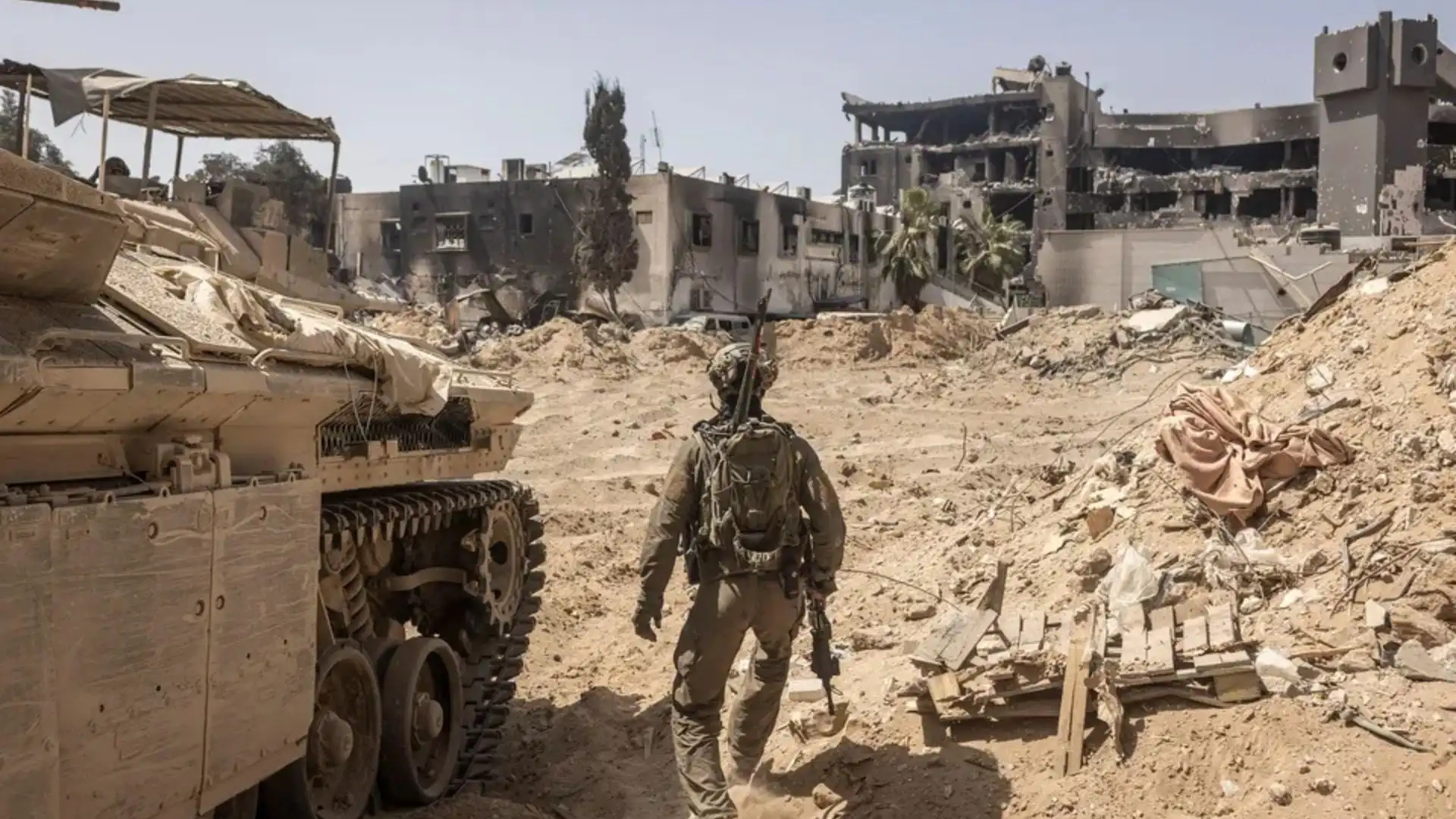The image shows a soldier walking through a devastated area, likely a combat zone. To his left, there is a military armored vehicle covered in dust, suggesting it has seen action. In the background, buildings are severely damaged, with partially destroyed structures and broken windows. The scene has an atmosphere of desolation, with debris scattered on the ground and dry air, giving the impression of a city or area ravaged by recent conflict. The soldier, armed and dressed in combat gear, moves through the wreckage, likely on a reconnaissance or security mission in the area.
