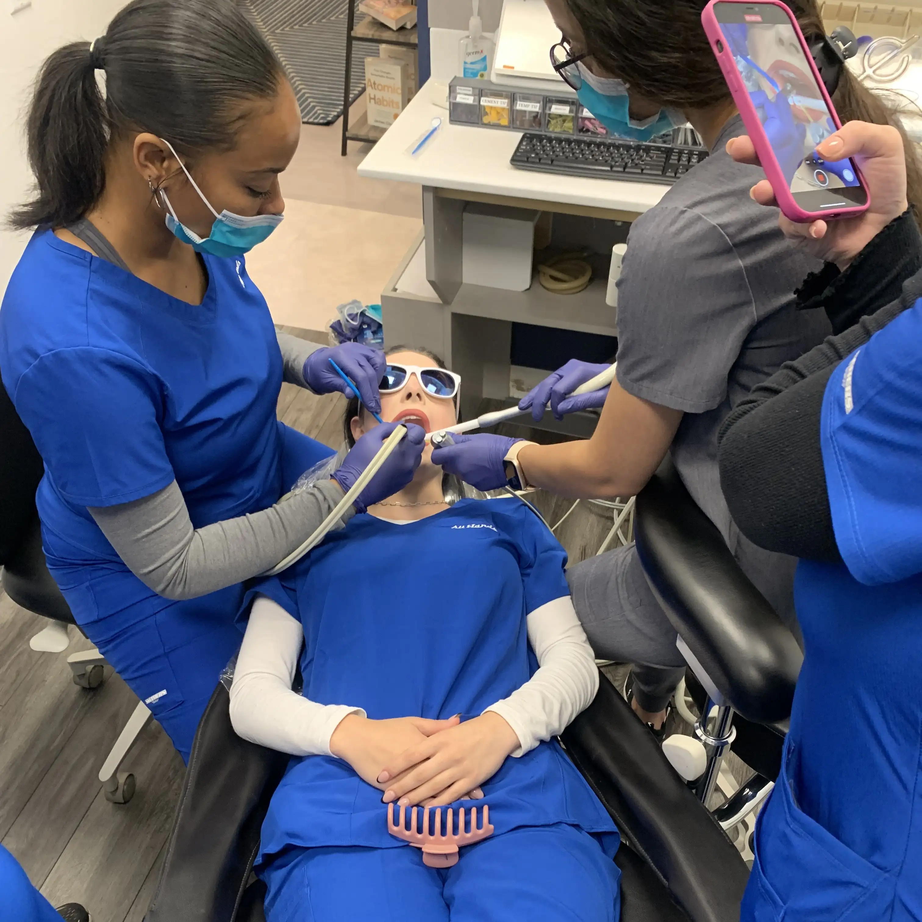 The image depicts a dental procedure being performed in what appears to be a clinic or training environment. A patient is seated in a dental chair, wearing protective glasses, while multiple dental professionals in blue scrubs use various instruments to perform the procedure. One individual is recording the process with a mobile device, suggesting that the session may be part of a training or educational program. The setting is a clean, organized dental office.